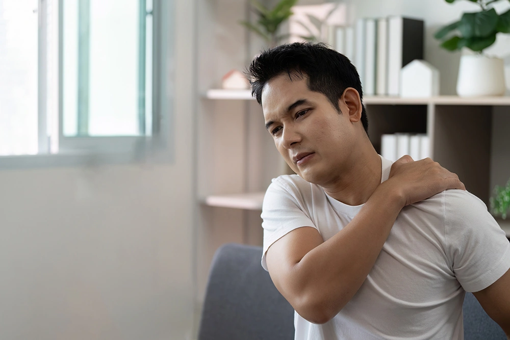 A man seated, gripping his shoulder experiencing pain in the area.