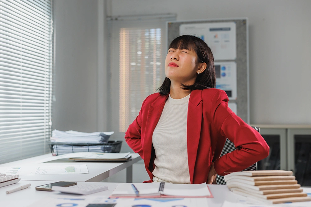 Woman seated at an office desk experiencing hip pain linked to prolonged desk-bound work.